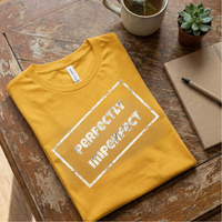 Load image into Gallery viewer, Mustard Yellow / Gold t-shirt flat lay on rustic wood. Features the white "Perfectly Imperfect" stamp design.