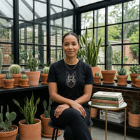 Load image into Gallery viewer, A serene woman seated in a sunlit botanical greenhouse, wearing a side-seamed, true black premium luxury tee. The high-contrast garment displays a vintage-inspired white line-art rabbit with gold mechanical glasses, placed next to books on resilience and botanical drawing.
