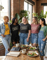 Load image into Gallery viewer, Group of four women laughing in a coffee shop, wearing "Perfectly Imperfect" t-shirts in Mustard Yellow, Black, Mauve, and Denim. Represents friendship and inclusivity.