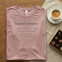Load image into Gallery viewer, Vital Threads "Hopeful Romantic" t-shirt in Mauve Pink. Flat lay on a wooden table with a cup of coffee and a box of chocolates. The graphic features a dictionary-style definition about finding beauty in the ordinary.