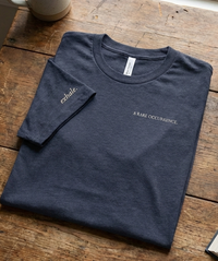 Load image into Gallery viewer, Flat lay photography of the women's premium sueded cotton t-shirt in Navy Heather. Features minimalist chest text 'A RARE OCCURRENCE' and 'exhale.' on the sleeve. Quiet luxury wellness apparel on a wooden surface.