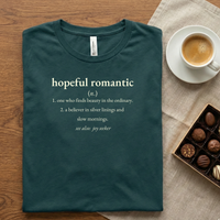 Load image into Gallery viewer, Forest Green definition t-shirt folded neatly on a wooden desk with espresso and chocolates. Features the "Hopeful Romantic" white text graphic.