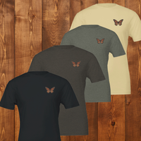 Load image into Gallery viewer, No. 26: Chrysalis to Flight - Stacked premium luxury tees in charcoal, forest green, and soft cream, each featuring the subtle botanical butterfly chest graphic.