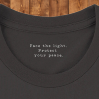Load image into Gallery viewer, Inner Collar Vintage Black: Close-up macro of the inner collar of the Vintage Black No. 28 The Evolution Premium Luxury Tee, showing the hidden somatic anchor text: Face the light. Protect your peace.