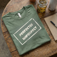 Load image into Gallery viewer, Olive Green t-shirt with "Perfectly Imperfect" box logo. Minimalist flat lay with coffee and a notebook.