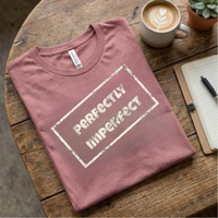 Load image into Gallery viewer, Mauve Pink graphic tee folded on a wooden surface. Highlights the "Perfectly Imperfect" text in a vintage stamp style.