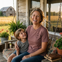Load image into Gallery viewer, Woman and child on a porch swing, woman wearing the Land of the Free, Home of the Brave shirt in Heather Mauve with the front 1776 star-circle badge visible.