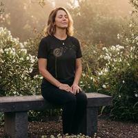 Load image into Gallery viewer, No. 25: The Emergence - Woman sitting peacefully in a garden wearing a high-contrast black premium luxury tee featuring a vintage botanical dandelion graphic