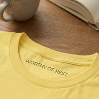 Load image into Gallery viewer, Close-up of the inner neck label on a yellow Vital Threads t-shirt reading "WORTHY OF REST" in minimalist font.