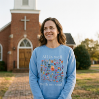 Load image into Gallery viewer, Woman standing outside a small brick church wearing the Carolina Blue All Is Well crewneck sweatshirt, the wildflower field and “All is well with my soul” visible across the chest.