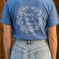 Load image into Gallery viewer, Back view of True Royal Blue t-shirt with white "Grace, Joy, &amp; Peace" floral wreath design. Modeled in a casual lifestyle setting.