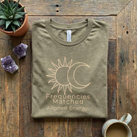 Load image into Gallery viewer, Olive Green spiritual wellness t-shirt folded on rustic wood with crystals. Features the "Frequencies Matched, Aligned Energy" graphic.