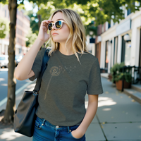 Load image into Gallery viewer, No. 25: The Emergence - Woman walking down a city street wearing an olive green premium luxury tee with a botanical dandelion graphic