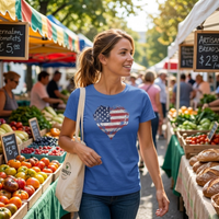 Load image into Gallery viewer, Woman walking through an outdoor farmers market wearing the Heather Columbia Blue Deep Roots, Steady Heart shirt with a distressed American flag heart graphic across the chest, market stalls and a canvas tote bag in view.
