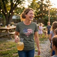 Load image into Gallery viewer, Woman laughing while carrying a pitcher of lemonade at an evening backyard gathering, wearing the Heather Military Green Deep Roots, Steady Heart shirt with a distressed American flag heart graphic across the chest, string lights and picnic tables in the background.