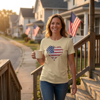 Load image into Gallery viewer, Woman walking down porch steps holding a coffee mug at sunrise, wearing the Soft Cream Deep Roots, Steady Heart shirt with a distressed American flag heart graphic across the chest, small-town street with flags on porches in the background.