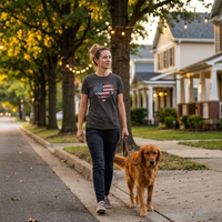 Load image into Gallery viewer, Woman walking a golden retriever on a tree-lined neighborhood sidewalk at golden hour, wearing the Vintage Black Deep Roots, Steady Heart shirt with a distressed American flag heart graphic across the chest.