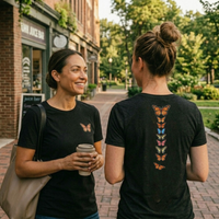 Load image into Gallery viewer, No. 26: Chrysalis to Flight - Two women conversing outdoors wearing premium luxury tees; left shows the subtle chest butterfly graphic, right shows the striking vintage botanical butterfly spine back print on charcoal.