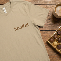 Load image into Gallery viewer, Soft Cream/Beige t-shirt featuring "Soulful" typography. Styled with chocolates for a warm, cozy vibe.