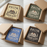 Load image into Gallery viewer, All four colorways of the Faith Can Move Mountains shirt — Soft Cream, Heather Military Green, Heather Columbia Blue, and Vintage Black — folded in open kraft gift boxes with tissue paper.