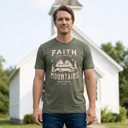 Man standing outside a small white church with a steeple, wearing the Heather Military Green Faith Can Move Mountains shirt with the Matthew 17:20 mountain graphic visible.