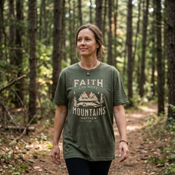 Woman walking through a pine forest wearing the Heather Military Green Faith can Move Mountains shirt, featuring the Matthew 17:20 mountain graphic across the chest.