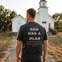 Load image into Gallery viewer, Man wearing Dark Grey Heather God Has A Plan t-shirt facing a small white church at golden hour, back print visible