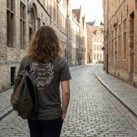 Load image into Gallery viewer, Lifestyle shot from behind of a woman walking down a cobblestone street wearing the Grace, Joy &amp; Peace shirt in Charcoal Grey, showing the back botanical wreath graphic with 'Grace, Joy &amp; Peace' and 'The Inner Work' text.