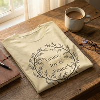 Load image into Gallery viewer, Back view of the Grace, Joy and Peace shirt in Soft Cream, folded on a wooden desk with coffee and glasses — botanical wreath chest graphic visible.