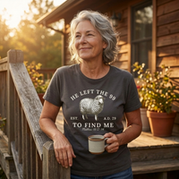 Load image into Gallery viewer, Woman holding a coffee mug on a porch at sunset wearing the He Left the 99 to Find Me shirt in Dark Grey Heather, front sheep graphic visible.