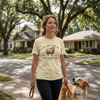 Load image into Gallery viewer, Woman walking her dog through a tree-lined neighborhood wearing the He Left the 99 to Find Me shirt in Warm Cream, featuring the arched sheep graphic and Matthew 18:12-14 reference.