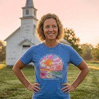 Load image into Gallery viewer, Woman standing in front of a white country church at sunrise, wearing the Heather Columbia Blue His Mercies Are New Every Morning shirt with the painted sunrise arch graphic and Lamentations 3:22-23 visible on her chest.