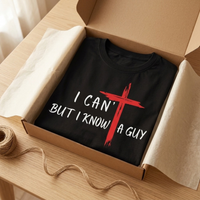 Load image into Gallery viewer, Black I Can't But I Know A Guy t-shirt folded in an open kraft gift box with cream tissue paper and twine, the red cross and white lettering facing up.
