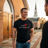 Load image into Gallery viewer, Man outside a brick church wearing the Black I Can't But I Know A Guy t-shirt, white hand-brushed lettering, I CAN' over BUT I KNOW A GUY, with a red brushstroke cross that forms the T in CAN'T and stands between KNOW and A GUY.
