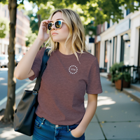 Load image into Gallery viewer, Woman wearing the Land of the Free, Home of the Brave shirt in Heather Mauve on a city sidewalk, front 1776 star-circle badge visible.