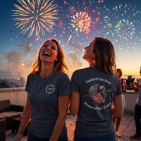 Load image into Gallery viewer, Two women in Heather Midnight Navy Land of the Free, Home of the Brave shirts watching fireworks at dusk — one showing the front 1776 badge, the other the full back eagle and flag graphic.