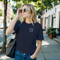 Load image into Gallery viewer, Woman wearing the Land of the Free, Home of the Brave shirt in Heather Midnight Navy on a city sidewalk, front 1776 star-circle badge visible.