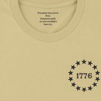 Load image into Gallery viewer, Collar detail of the Land of the Free, Home of the Brave shirt in Warm Cream, showing the hidden inner collar message ‘Freedom was never free. Someone paid so you wouldn't have to.’ above the front 1776 star-circle badge.