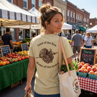Load image into Gallery viewer, Woman at a farmers market wearing the Land of the Free, Home of the Brave shirt in Warm Cream, back eagle and flag graphic with arched banner text visible in Deep Espresso ink.