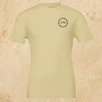 Load image into Gallery viewer, Front flat lay of the Land of the Free, Home of the Brave shirt in Warm Cream, showing the 1776 star-circle badge in Deep Espresso ink and inner collar message at the neckline.