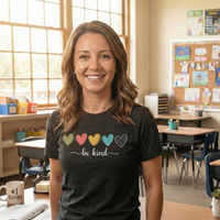 Load image into Gallery viewer, Smiling female teacher in a classroom setting wearing the charcoal black 'Everyday Kindness' premium graphic tee. Features five colorful hearts and the script text 'be kind'. High-quality apparel for educators and mindful women.
