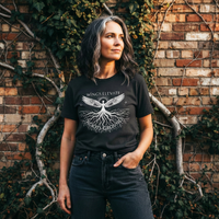 Load image into Gallery viewer, A woman in an architectural setting wearing the No. 21 Equilibrium botanical tee featuring celestial wings and grounded roots.