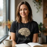 Load image into Gallery viewer, A lifestyle photograph of a smiling woman with shoulder-length brown hair and a micro cross necklace, sitting in a modern cafe and making direct eye contact with the viewer. She is wearing the premium Heather Black "Hold On Let Me Overthink This" graphic t-shirt by Vital Threads Clothing.
