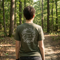 Load image into Gallery viewer, Outdoor lifestyle shot of a person walking in the woods wearing the Heather Military Green No. 11 The Trilogy premium luxury tee, showcasing the back botanical wreath design.