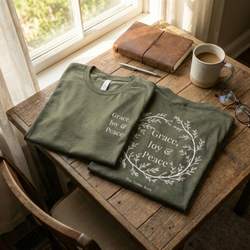 Flat lay on a rustic wooden table featuring two Heather Military Green No. 11 The Trilogy premium luxury tees, showcasing the front chest typography and back botanical wreath graphic.