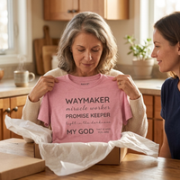 Load image into Gallery viewer, A woman receiving the Soft Pink Waymaker, Promise Keeper shirt as a gift, unwrapped from a box at a kitchen table while a younger family member looks on.
