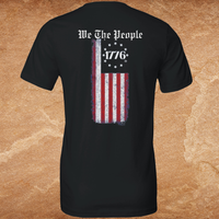 Load image into Gallery viewer, Flat lay back view of the We The People premium tee in black showing the full We The People gothic blackletter print and distressed vertical American flag graphic with 1776 detail