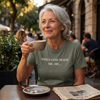 Load image into Gallery viewer, Silver-haired woman drinking coffee at an outdoor café table wearing the When God Made Me shirt in Heather Military Green, front "When God Made Me, He..." text visible.