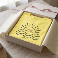 Load image into Gallery viewer, The Yellow You Are The Light shirt folded in an open gift box with a ribbon and tissue paper on a pale wood table, the collar message visible at the neckline.
