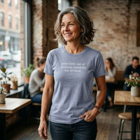 Load image into Gallery viewer, Silver-haired woman in a warm cafe wearing the Your Little Ray of Sarcastic Sunshine Has Arrived shirt in Heather Blue, white script lettering on the front.